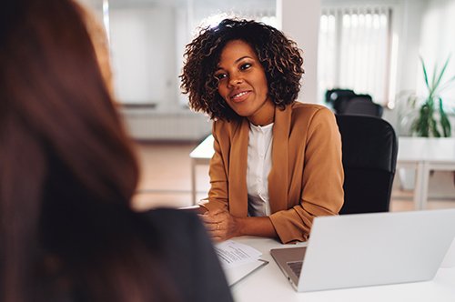 a Black woman in professional attire leans forward toward to speak to another woman
